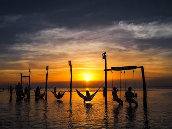 Silhouette people resting on swings and hammocks over sea against sky at sunset