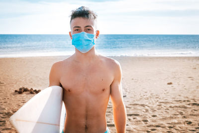 Portrait of shirtless boy wearing mask holding surfboard standing on beach