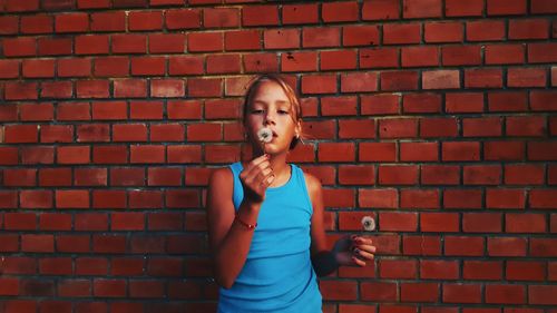 Girl blowing dandelion while standing against brick wall