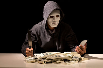 Criminal in mask using mobile phone while sitting with currency at table against black background