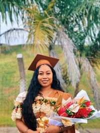 Portrait of smiling young woman in graduation gown standing against trees