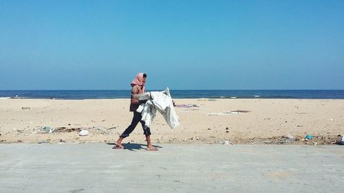 Side view of man holding sack while walking at beach against clear blue sky