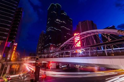 Light trails on road by buildings against sky at night