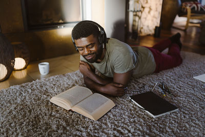 Smiling young man listening to music and reading book while lying on carpet at home