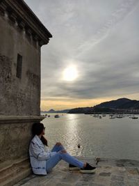 Woman sitting by sea against sky during sunset