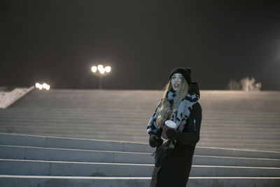 Portrait of woman holding coffee while standing on steps against sky at night