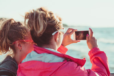 Rear view of mother photographing sea while standing with daughter against sky during sunset