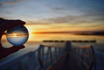 Cropped hand of person holding crystal ball against sky during sunset