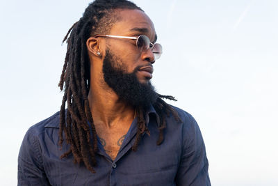Black male with dreadlocks in sunglasses standing on coast and looking away against gray sky