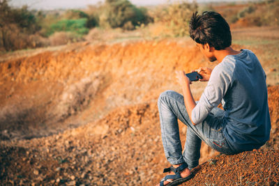 Side view of young man using mobile phone while sitting on rock