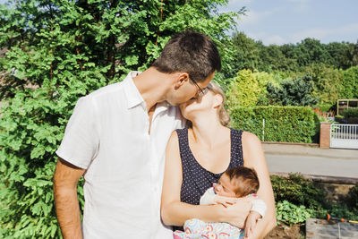 Woman holding baby kissing man while standing against trees