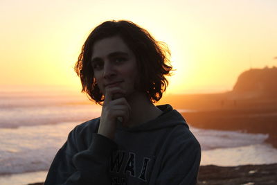 Portrait of young man with hand on chin against beach during sunset