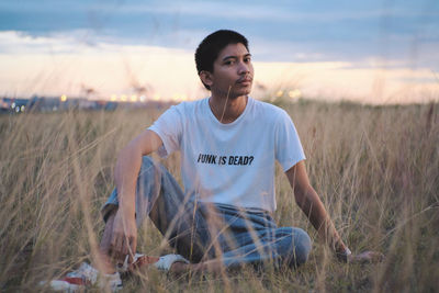 Portrait of young man sitting on grassy land against sky at sunset