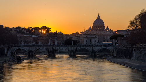 Bridge over river by buildings against sky during sunset