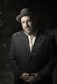 Portrait of well-dressed man wearing hat standing outdoors at night during snowfall