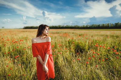 A girl in a red dress above the knee stands in a poppy field during the day against the sky