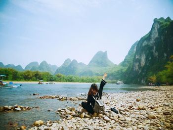 Woman gesturing peace sign while sitting at lakeshore against mountains