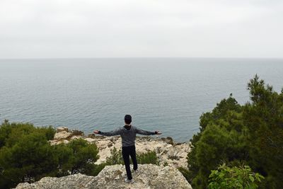 Rear view of man with arms outstretched standing on cliff against sea