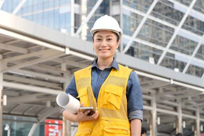 Portrait of mid adult woman holding mobile phone and blueprint while standing against building