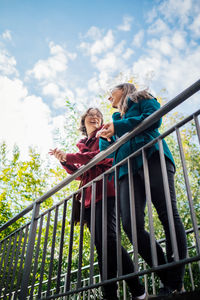 Low angle view of cheerful sisters standing by railing on bridge against sky