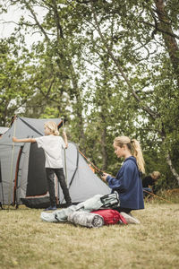 Teenage girl using phone while family pitching tent at camping site