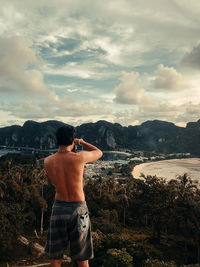 Rear view of shirtless man photographing water against sky