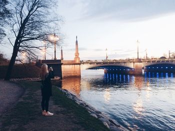 Full length of woman photographing illuminated bridge using smart phone while standing at riverbank during sunset