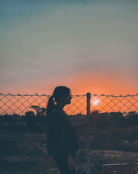 Side view of woman standing by chainlink fence against sky during sunset
