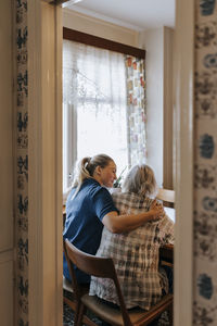 Female caregiver with arm around of senior woman sitting on chair at home