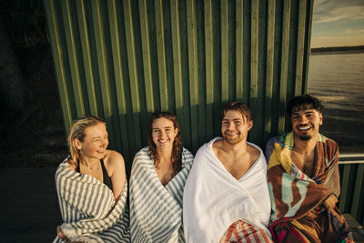 Portrait of happy male and female friends wearing towel while sitting against cottage