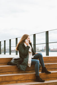 Young woman using mobile phone while sitting on seat against sky