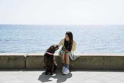 Woman sitting with labrador dog on retaining wall in front of sea