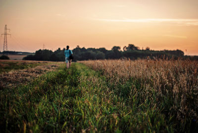 Rear view of woman walking on grassy field against sky during sunset