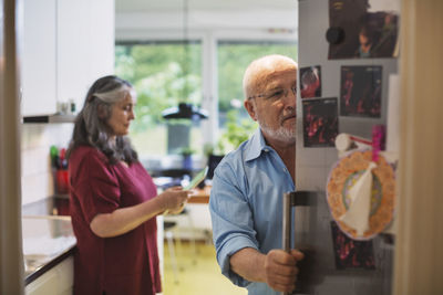 Senior man opening refrigerator while woman using digital tablet at home