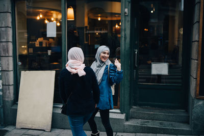 Happy young woman waving while walking with friend on sidewalk in city