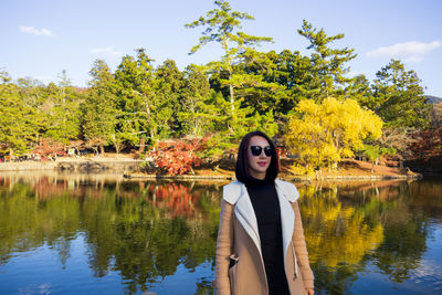 Portrait of young woman standing by lake against trees