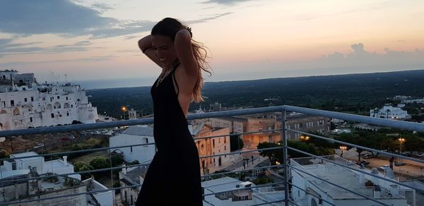 Woman standing by railing against buildings during sunset