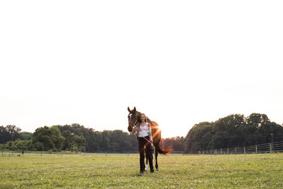 Camera aware teenage girl stands with brown horse in field at sunset