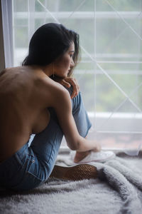 Shirtless young woman sitting by window at home