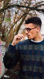 Young man smoking cigarette while standing against tree