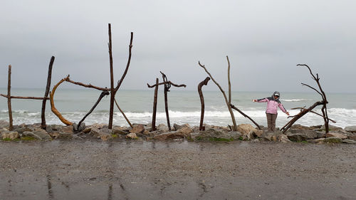 Woman with arms outstretched standing amidst driftwood at beach against sky