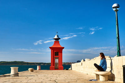 Side view of woman sitting on seat at pier against lighthouse