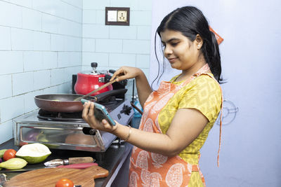 A pretty indian young woman wearing apron watching cooking video in smart phone in domestic kitchen