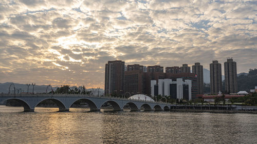 Bridge over river by buildings against sky during sunset