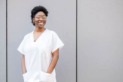 Delighted african american female in white wear and glasses and work uniform looking at camera with smile while standing on street near wall