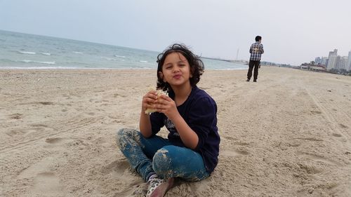 Girl having food while sitting at beach against sky