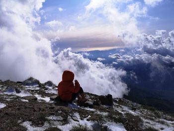 Rear view of kid sitting on rock against sky during winter