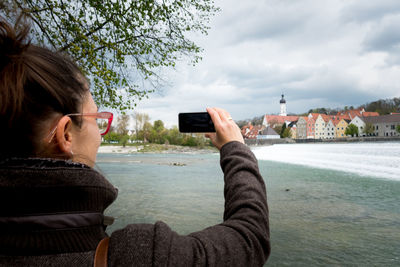 Rear view of woman photographing cityscape by lech river against cloudy sky