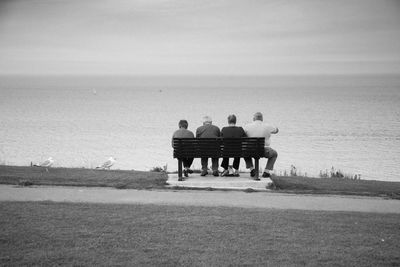 Rear view of men sitting on bench by sea against sky