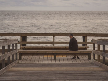 Man sitting on bench by pier at sea against sky